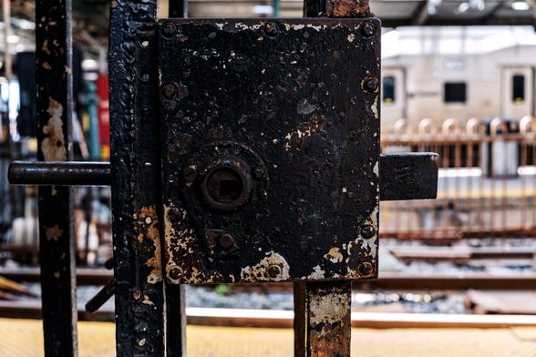 A color photograph taken during daytime focusing on the chipped black paint on a large, square latch on a train station platform gate. The bolt extends out past the right edge of the plate.

On the right, tracks and platforms are visible, defocused, and a silver train sits several tracks back. To the left edge, visual clutter at the far end of the platforms, some red, some green, with whites and browns stands in jumble.