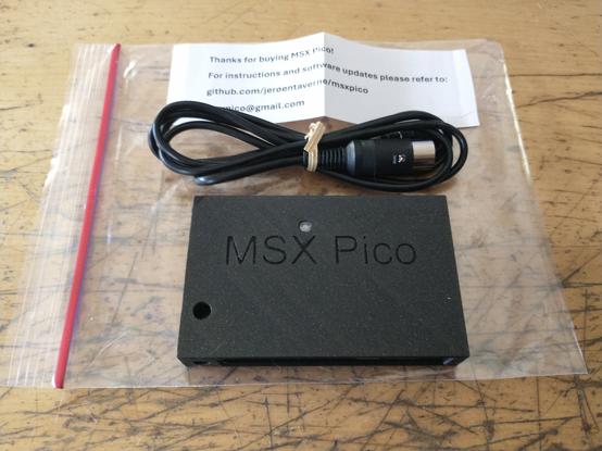 A black "MSX Pico" cartridge and a MIDI Cable for that cartridge resting on the transparent plastic bag they were packed in.