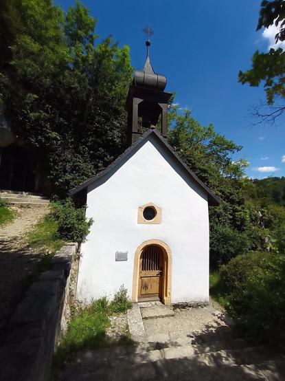 Vue en léger surplomb, chapelle à la façade blanche et clocheton bizarrement galbé.
En arrière plan, forêt en pente et ciel bleu profond.