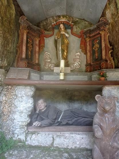 Dans le fond d'un abri sous roche et dans une niche, un moine en bois est allongé sur le côté. Il s'est à demi redressé, accoudé, la joue appuyée sur  la main.
Au-dessus, un autel coloré où trône une Vierge drapée de vert, or et argent 
Devant le moine, cachant ses pieds, un ours sculpté.