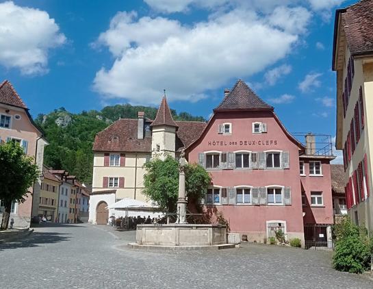 Une autre fontaine sur fond de maisons, dont une jaune pâle à tourelle, et un hôtel vieux rose.
Ciel bleu nuageux.