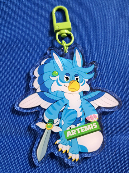 Photographic of a small acrylic charm, depicting an anthro blue female gryphic drawn in a chibi style, with a confident expression while wielding a a sword. The charm has a green clasp and a green rectangle name tag that reads "Artemis".