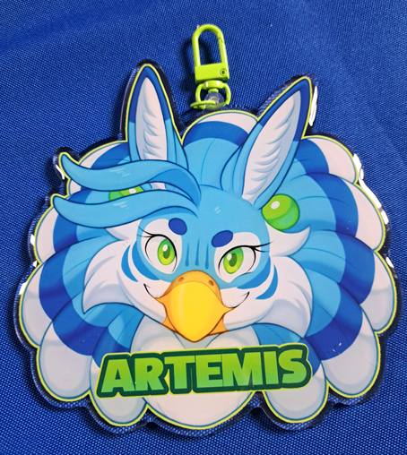 Photograph of a large acrylic charm badge, with a bust illustration of a blue female gryphon looked forward with a confident smirk. The charm has a green clasp and has text below the character's face with her name, Artemis.