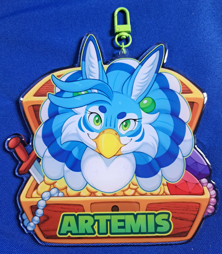 Photograph of a very large acrylic charm badge, with a bust illustration of a blue female gryphon looking towards the viewer with a confident smirk, and a backdrop depicting an open treasure chest with a sword, pearl strings, large gems and gold inside. The charm has a green clasp and text that reads "Artemis" on the front side of the chest below the drawing of the character.