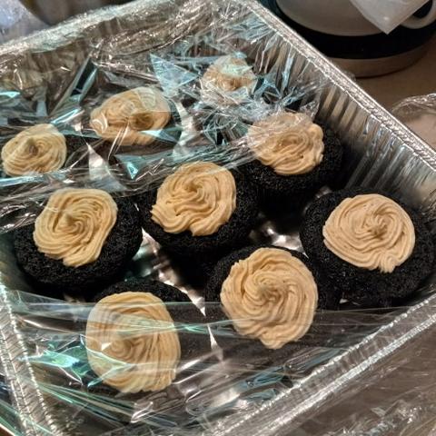 An aluminum pan with nine very very dark chocolate cupcakes with brown icing wrapped in cling wrap, sits on a counter with the cling pulled back a bit so you can se the cupcakes in all their delicious glory.