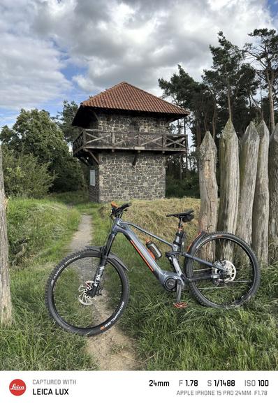 Der Limesturm bei Watzenborn-Steinberg, Gemeinde Pohlheim, Landkreis Gießen. Im Vordergrund lehnt ein eMTB an einer Palisade aus Baumstämmen. Zwischen den Baumstämmen für ein Weg zum Turm.