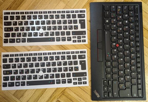 two sheets of keyboard stickers from tastatur-aufkleber.eu for NEOqwertz layout for Lenovo keyboards next to a keyboard where the stickers have been applied.