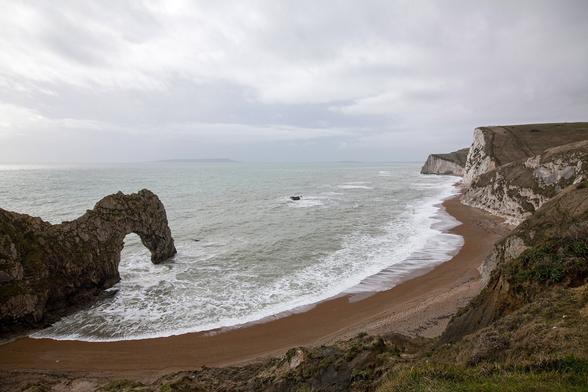 It's an overcast day with sheets of grey cloud overlapping all the way to the distant horizon that we can see from our vantage point on top of cliffs over the shoreline. To the right the cliffs continue around and out to sea, topped with grass and fronted by sheer faces of white chalk. Where we're standing it looks a little less like a straight drop in front of us with some of the grassy headland dipping down to where we can see a crescent shape of damp-looking sand curling around in front of the cliffs. The beach's border with the sea is a thick band of foam from the breaking waves but the sea looks relatively calm and grey-green from this distance and height. Emerging from land from the left of the view is a rocky promontory with a natural arch in it. It appears like a giant, rusted, old bottle opener jutting into the water.