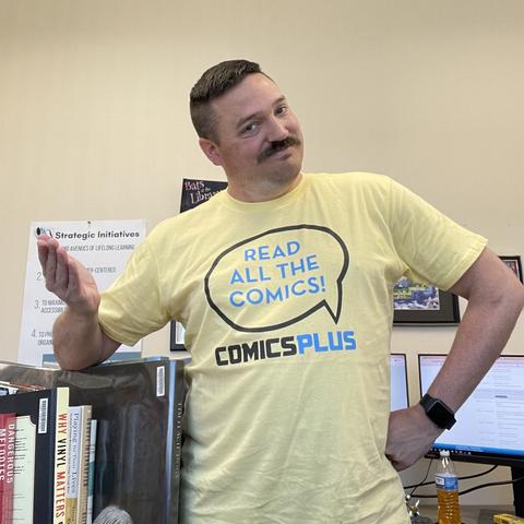 A person with a mustache is wearing a yellow "Comics Plus" T-shirt with a speech bubble saying "Read all the comics!" They are standing with one hand on their hip and the other gesturing to the side. There are books and