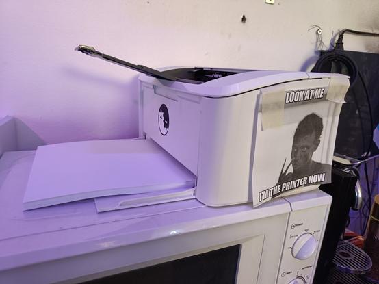 A small monochrome printer on top of a microwave oven. On the side is a picture of the "Look at me now" meme with the text "Look at me -- I'm the printer now"