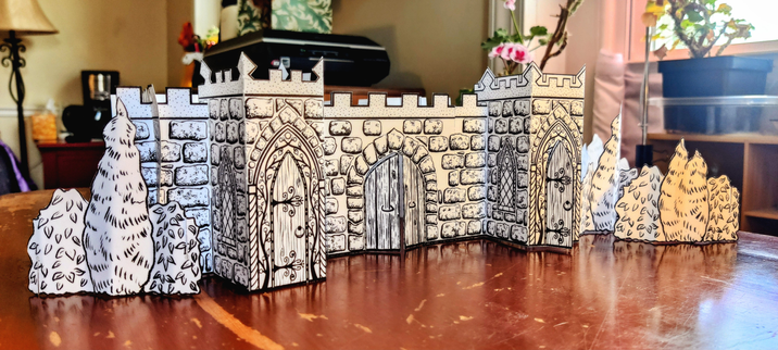 A photo of the front of a 3D paper castle made of sections of white paper printed with stones, crenellation, doors, and windows in black ink. The castle angles away to the right. The front wall of the castle has a vestibule on either side, each with a door in front with a pointed gothic arch, surrounded by decorative vines with a tiny sun at the top. Each vestibule also has windows on either side, with pointed gothic arches and stained glass. In the center of the front wall, between the vestibules, is a larger but simpler double wooden door with a gothic arch, standing slightly ajar. Paper shrubbery stands to either side of the castle. The castle sits on a scratched wooden desk. A white and pink geranium is blooming behind and to the right of the castle.