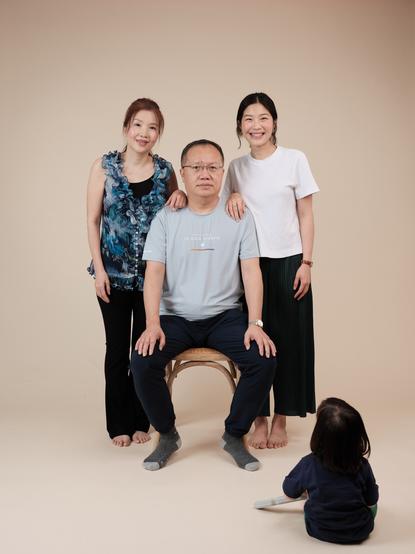 A studio portrait of a seated man, his wife and daughter sanding on either side of him, and his baby grand-daughter playing on the floor, facing away from the camera.