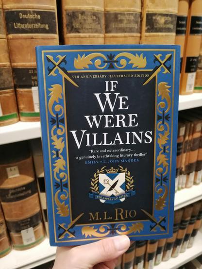 Holding the novel "If We Were Villains" in SUB Göttingen library, in front of bookshelves filled with old, leather-bound books