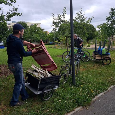Dans un petit verger urbain, 2 hommes commencent à décharger des remorques vélo remplies de matériel de jardin et cageot de jeunes pousses, sous un ciel nuageux