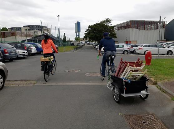 Femme casquée à la veste orange vif et homme en jeans et béret s'élancent sur des vélos chargés de plantes et matériel de jardin, dans une rue et parkings de banlieue remplis de voitures