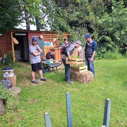 Dans un jardin avec une cabane en bois sous les arbres, des hommes s'apprêtent à transporter des cageots de jeunes pousses sous les visages souriants d'une homme et d'une femme