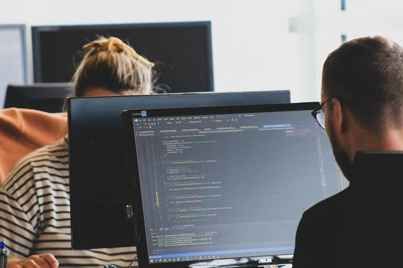 Photograph shows two programmers facing each other, sitting in front of their computer in a work space. Image by Rivage under a Free Unsplash License: https://bit.ly/3Ks304N.