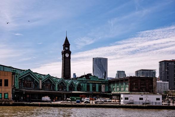 A color, daytime photograph of  Hoboken Terminal taken from Pier A Park. Water (the Hudson) sits between the pier and the terminal. The terminal extends from the left edge of the frame (the ferry terminals), angled away from the viewer, bends to the right (main lobby entrance) and again to form a shorter “wing” (the terminal end structure for the platforms and Bush-type train sheds). The center of the check-mark shape is a public square, which currently houses a white construction trailer. The entire facade is covered in copper, displaying a green/blue patina. A stone clock tower, a later reconstruction of the original, rises behind, lit letters stacked vertically to spell “Lackawanna”. Buildings in Hoboken are visible to the right and behind. A jet aircraft heading north and a helicopter heading southeast are visible in the sky on the left.