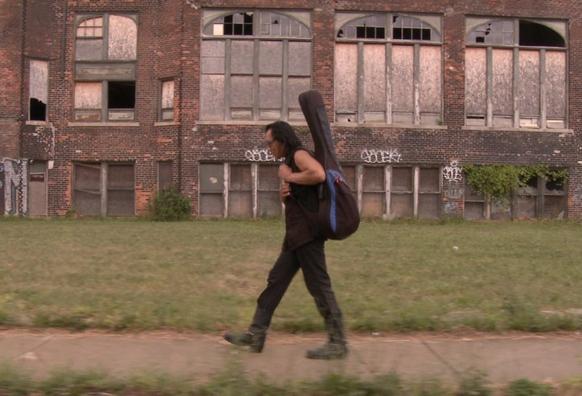 A photo of Sixto Rodriguez, with a guitar case on his back, walking through a working class neighbourhood in Detroit. There is a rundown building with broken windows behind him as he walks.