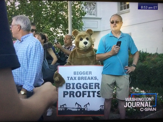 screen grab from cspan time stamped 853 am est of a person in a bear costume carrying a sign that says "bigger tax breaks, BIGGER PROFITS" with oil field imagery. They are outdoors w paps behind them, white guys in khakis and blue shirts stand on either side