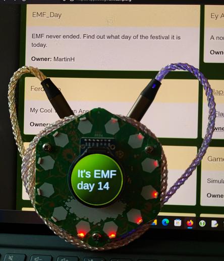 Photo of EMF Tildagon badge displaying the message "It's EMF day 14". In the background is the #Tildagon app store showing an entry for an app called EMF_Day. The description for the app says "EMF never ended. Find out what day of the festival it is today"