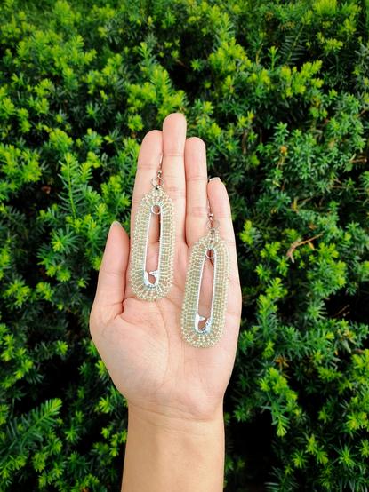 A pair of lightly green colored beaded safety pins being held by Kim's hand over a growing green shrub.