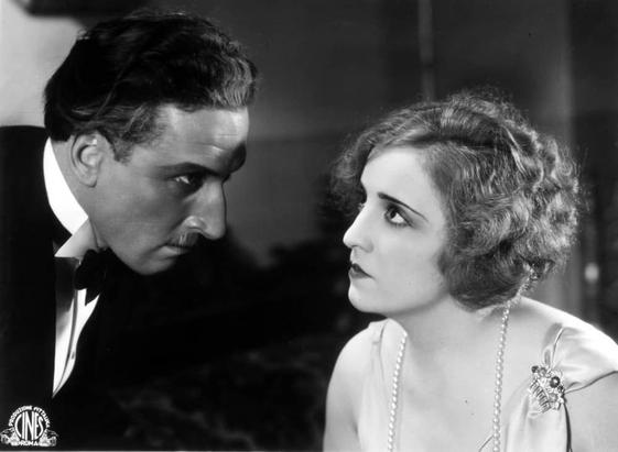 The image depicts two individuals engaged in a conversation. On the left side of the image, there is a man wearing formal attire, including what appears to be a suit and tie, suggesting an elegant or formal setting. On the right side, there is a woman with short hair, also dressed in evening wear that includes a sleeveless dress, which conveys a sense of formality consistent with the male's attire. Both individuals are looking at each other with serious expressions on their faces, indicating an intense or important discussion. The background is minimal, allowing for the focus to be on the interaction between the two characters.