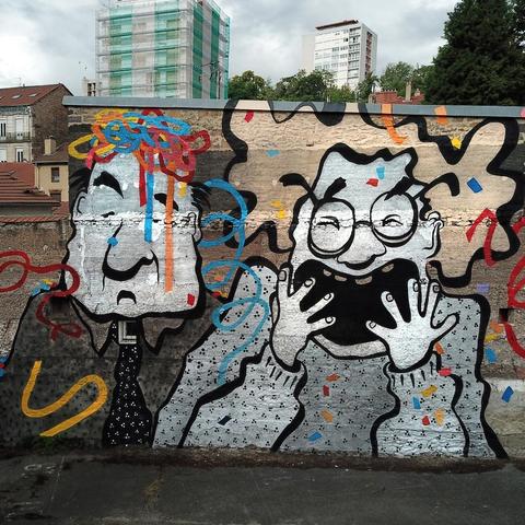 Streetartwall. A large paste-up with caricature drawings of two grotesque figures are displayed on a street wall. An elderly lady with blonde hair and glasses, dressed in a gray sweater, screams with her mouth open. She has raised her hands in front of her mouth in joy and colorful confetti and streamers are flying around her. Next to her is a gentleman in a gray suit and tie, on whose head some streamers have landed. He doesn't seem so enthusiastic.