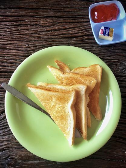 das hochformatige bild zeigt im unteren bereich einen hellgrünen frühstücksteller mit vier zu dreiecken halbierten scheiben toastbrot, die getoastet übereinandergefächert sind. außerdem ist auf dem teller ein frühstücksmesser. oben rechts im bild ist ein hellblaues schälchen mit zwei fächern. das obere ist gefüllt mit einer art roten marmelade, die sehr viel zucker enthält. im unteren der beiden fächer ist ein halbiertes stück butter, wie es sie in der hotelerie und gastronomie oft gibt, in alufolie verpackt. hier ist es in der umverpackung mit einem messer halbiert worden.
beides, teller und schälchen, stehen auf einem alten dunkelbraunen holztisch mit groben querlaufenden fasern.
für mein befinden geizte die betreiberin dieses kleinen hotels in der thailändischen stadt trat sehr mit der butter. zwei dieser kleinen butterstücke hätte ich angemessen gefunden. stattdessen gab es nur ein halbes.