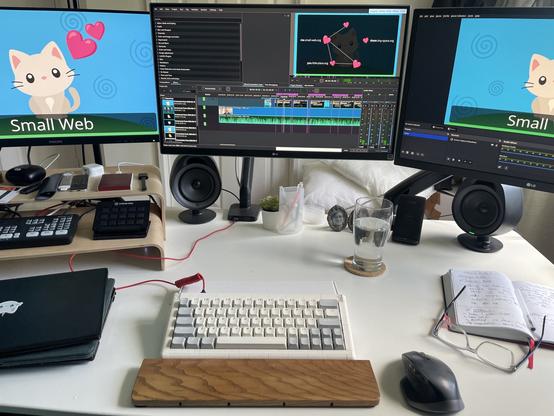 Photo of my desk with my presentation on slides.com open in the monitor on the left, showing the opening slide with an illustration of a minimalist grey kitten with pink ears and nose with the hearts emoji next to it and the title Small Web, Kdenlive non-linear editor open with the the timeline and another slide (showing places on the Small Web as hearts with domain names next to them – me.small-web.org, you.little-place.org, them.tiny-space.org – and four out of the five have some connections to one another denoted by dotted lines), and OBS open ready to screen capture the slides on the left monitor to be spliced into the original recording.

(There’s also a working LEGO keyboard, wooden wrist rest, a Logitech mouse, my diary/workbook, a pair of glasses, a glass of water, a stack of laptops, an Atem and Elgato Stream Deck and other miscellaneous gadgets on the desk.)