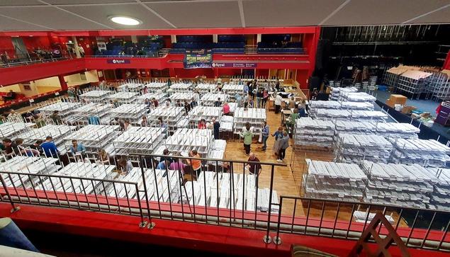 Widescreen photo, taken late morning on Sunday 9 June, looking down from the balcony at the European elections count centre in the TF Royal in Castlebar, Ireland. The space is dominated by rows of a couple dozen specially constructed open shelves, each containing pigeon holes for the 27 candidates. People work standing in the alleys between the shelves, and at desks along the entire perimeter. To the right are about 15 shelves containing thousands of still-unsorted ballot papers. Beyond them is the stage, where the results are read out and announcements made. Along the red balcony opposite and to the left are news reporters with TV and radio equipment.