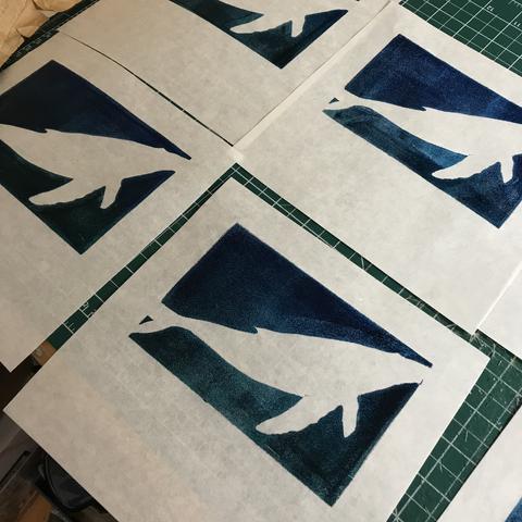 Prints laid out on a table with green cutting mat to dry. Parts of 6 square, 8” x 8” prints on white washi paper can be seen. The upper two thirds are printed as a deep blue-green rectangle with missing unprinted white silhouette of a humpback whale.