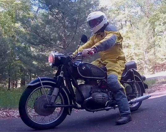 A person in a bright yellow and grey motorcycle suit, wearing a white helmet, astride an old black BMW motorcycle.