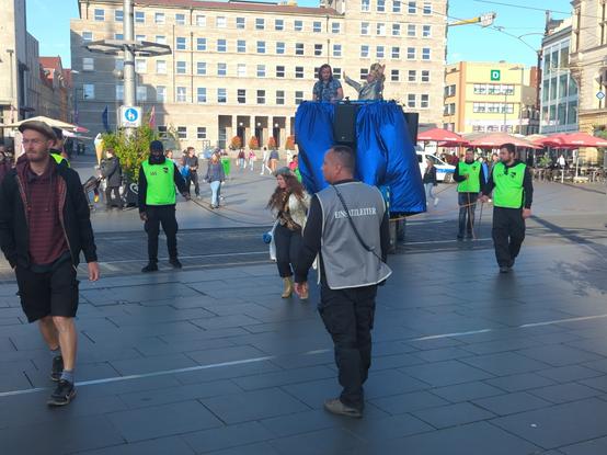 der König auf einem Selbstfahrer fährt über die Gleise der Straßenbahn und "befragt" seine Untertanen.
es laufen Lilliput mit, gesichert von Ordnern und Personal.
