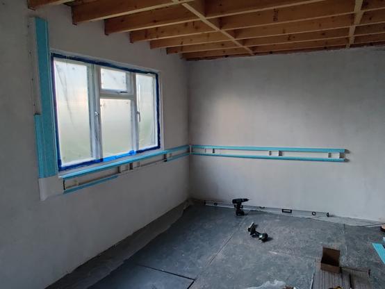 inside view of the front and right wall of the office construction. the walls are white and there is a large window on the front wall. there is dado trunking on both walls. empty backboxes are in the trunking and blue protective plastic covers exposed faces.