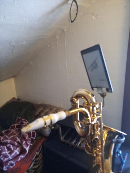 An attic room with a sloped ceiling, a cot and brown leather trunk in the background.  In the foreground is the upper portion of a baritone saxophone on a stand with a white mouthpiece.  Attached to the saxophone is a tarnished brass music clip holding a small ereader tablet.  Masking tape can be seen wrapping padding added to the metal clip in order to protect the tablet from damage.
