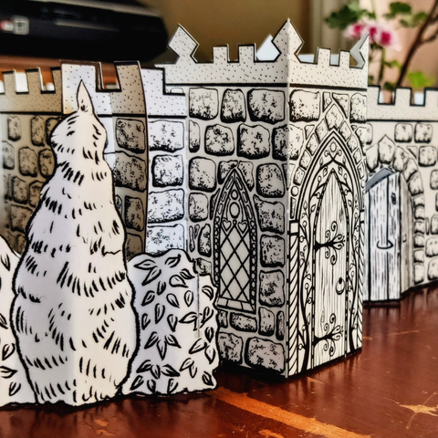 A closeup photo of Castle Vestibule 2.0 on the front left side of a 3D paper castle. The castle is made of sections of white paper printed with stones, crenellation, doors, and windows in black ink. At the front of the vestibule is a door with a pointed gothic arch, surrounded by decorative vines with a tiny sun at the top. Part of another arched wooden door is visible on the wall to the right of the vestibule. Paper shrubs stand to the left of the castle. The blurred green leaves of a geranium are visible in the top right of the photo.