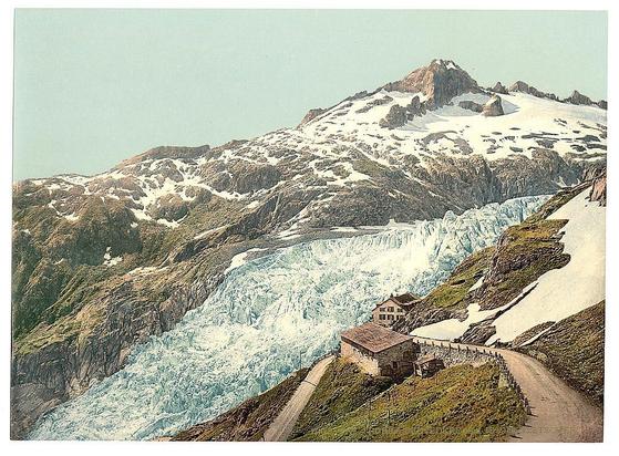 This is a vintage postcard depicting the Rhöne Glacier in Switzerland. The image captures the grandeur of the glacier, with its white surface contrasting against the lush greenery and mountains that surround it. A road can be seen near the base of the glacier, indicating human activity close to this natural wonder. The art style is characteristic of postcards from the late 19th or early 20th century, with a focus on capturing the beauty of nature at that time.