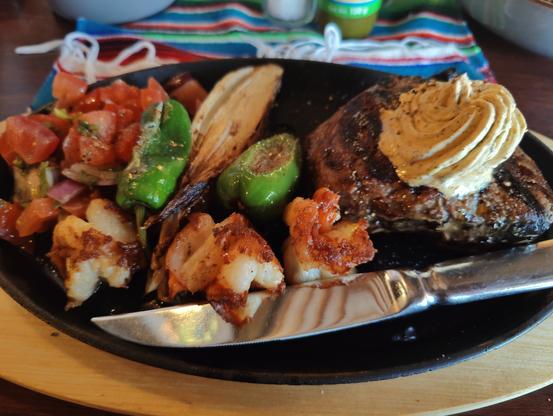 Ein Teller mit einem großen Steak mit hausgemachter Kräuterbutter darauf, daneben gegrillte Garnelen, Pico de Gallo Tomatensalat und gegrillten Peperones
