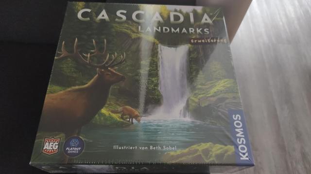Die Box der Brettspiele Erweiterung Cascadia Landmarks auf der Couch eines Hotelzimmers.