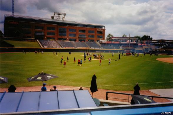 Farbfoto - ein Teil des Baseball-Stadions der Durham Bulls in Durham, North Carolina, USA. In der Bildmitte grüner Rasen und Leute, im Hintergrund Teile der Zuschauertribüne, dahinter ein großes braunes Backsteingebäude. Bewölkter Himmel. Kodak Ektar-Film - stark gesättigte Farben.