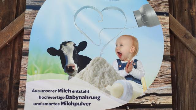 Bild eines Werbeplakats an einer Scheune: Eine Kuh und ein Baby neben einem Haufen Milchpulver; darüber eine Milchkanne, die Milch in Herzform ausleert und in diesem Haufen Milchpulver ankommt. Darunter der Text: "Aus unserer Milch entsteht hochwertige Babynahrung und smartes Milchpulver.