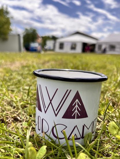 Weiße Emaille Tasse mit lila VVoid Logo steht in der Wiese bei Sonnenschein.