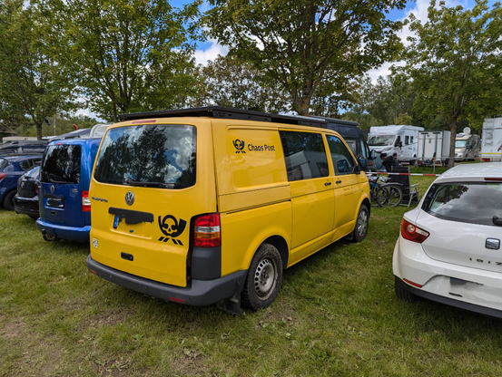 Gelber VW Transporter mit Chaos Post Logo hinten unter vorne. Das Fahrzeug ist auf einer Wiese zwischen anderen Fahrzeugen geparkt.