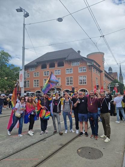 Das Foto zeigt Menschen welche in einer Reihe posieren vor eine rote Haus auf Tramgeleisen. Einige mit erhobener Faust, sie tragen bunte Kleidung slsie die Progressive Oride Flaggen von #kapers ubd des #sgbguss