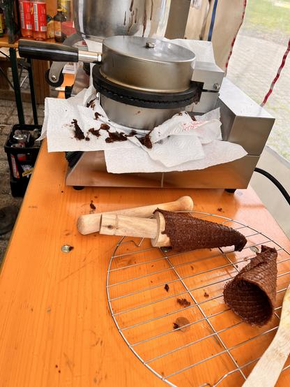 Es liegen braune Waffeln um ein kegelförmiges Holz herumgewickelt neben einem Waffeleisen. Die braunen Waffeln formen sich dadurch zu Eistüten