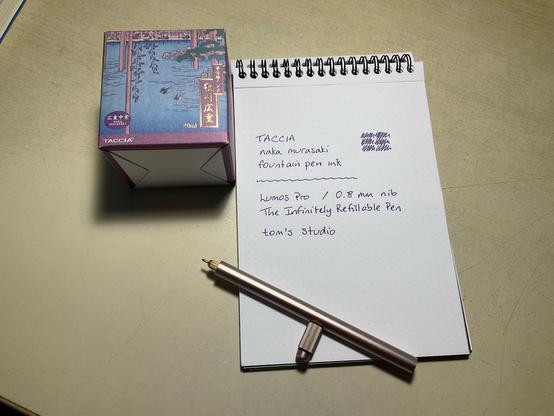 Ink swatch of TACCIA purple ink on a Rhodia A5 dotpad. Beside the pad is the naka murasaki ink box from TACCIA and beneath the swatch is the Lumos refillable pen in rose gold.