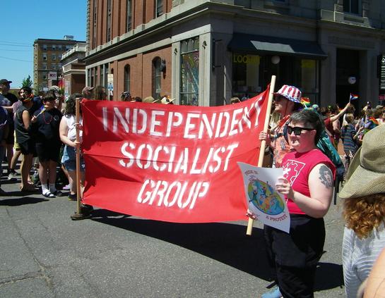 People at a Pride parade, holding up a red banner with the words, "INDEPENDENT SOCIALIST GROUP" written in white.