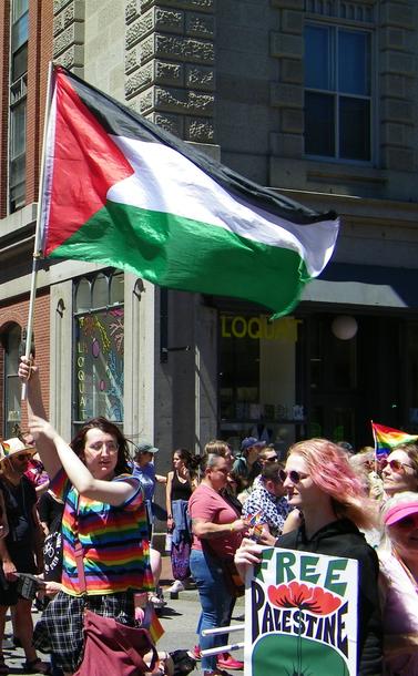 Marchers at a Pride Parade, one holding a large Palestinian flag, the other holding a sign reading, "FREE PALESTINE".