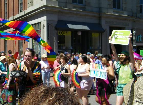 People marching in a Pride parade, many of them holding rainbow flags or fans. One women is holding a sign that reads, "QUEERS FOR CLIMATE JUSTICE," while the man next to her, who is wearing green, is holding up a sign that reads, "PROTECT FORESTS IN MAINE".